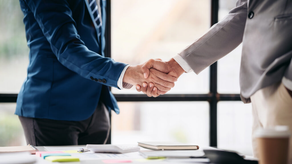 Zwei Personen in Businesskleidung besiegeln eine Zusammenarbeit per Handschlag über einem Schreibtisch.