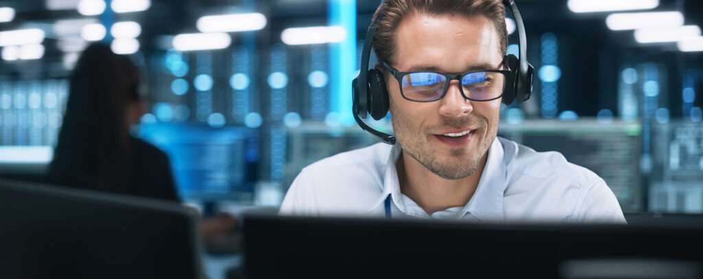 IT-Support-Mitarbeiter mit Headset und Brille arbeitet lächelnd am Bildschirm in einem Rechenzentrum.