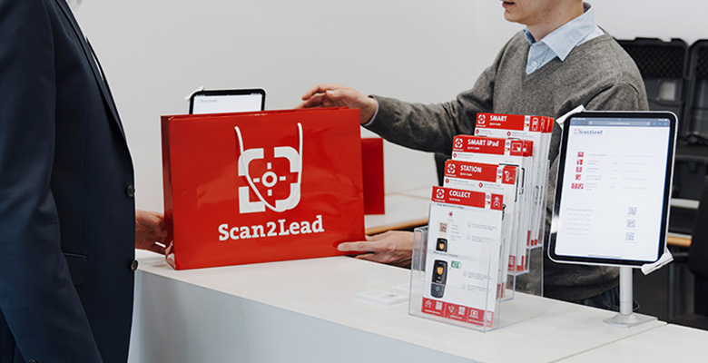 Mitarbeiter reicht am Service-Counter eine rote Scan2Lead-Tasche über den Tresen, daneben Info-Material und ein Tablet – technischer Support vor, während und nach der Messe.
