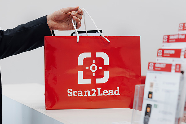Hand hält eine rote Scan2Lead-Tasche über einen Messe-Counter mit Info-Material – Abholung von Scanner und Einweisung am Service-Counter.