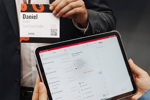 Person hält ein Leih-iPad mit geöffneter Scan2Lead App SMART und Lead-Ansicht, im Hintergrund ein Besucher mit Badge – Badges und Visitenkarten scannen.