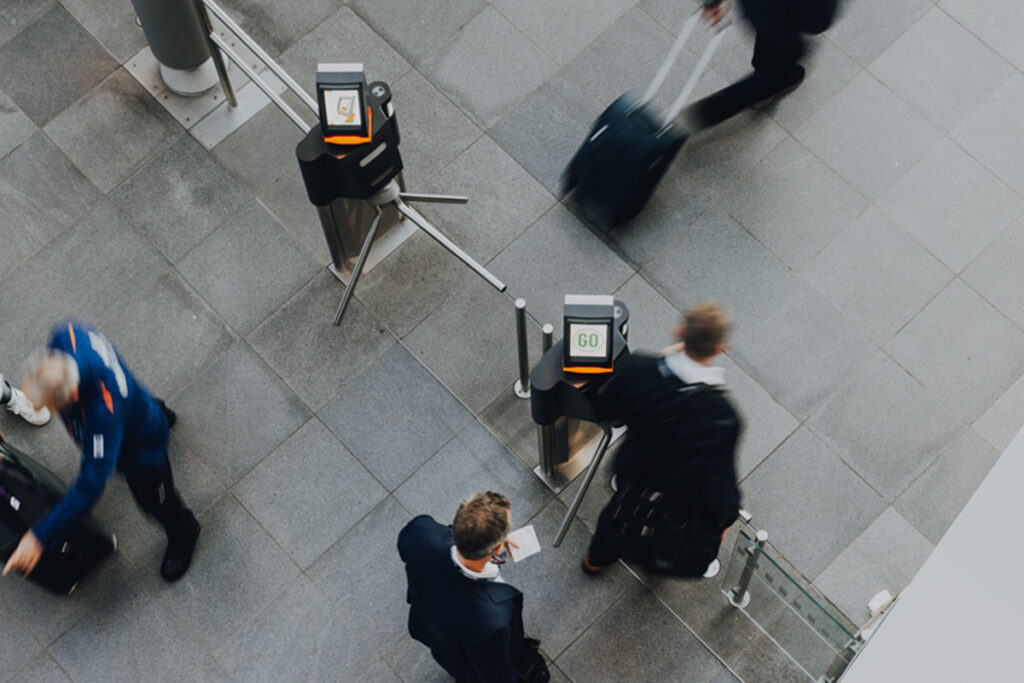 Blick von oben auf Drehkreuze mit Scan-Displays am Eingang einer Messe – Besucher passieren die Zugangskontrolle über ihr Badge, während im Hintergrund weitere Personen eintreffen.