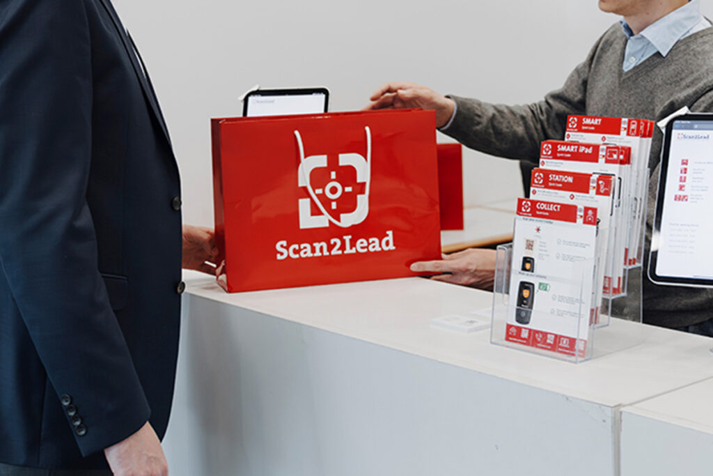 Mitarbeiter überreicht an einem Scan2Lead Service-Counter eine rote Scan2Lead-Tasche an einen Aussteller.