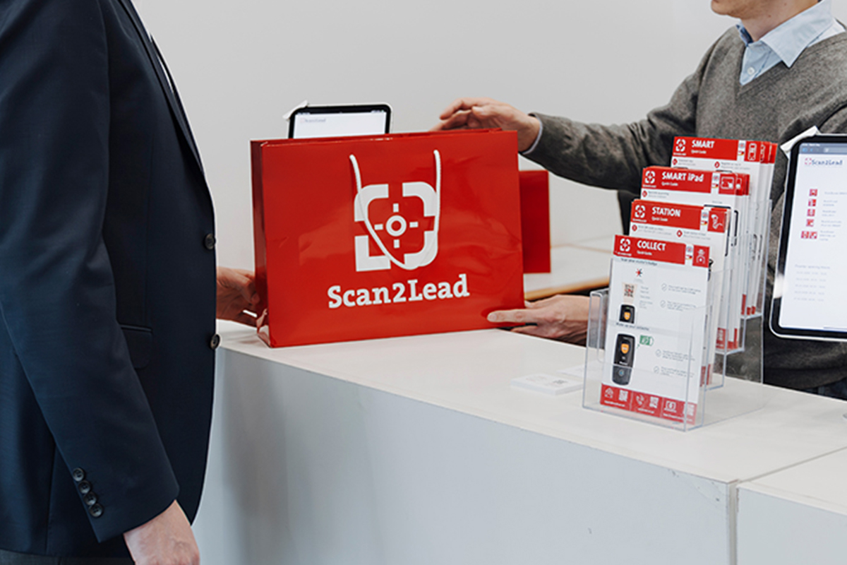 Scan2Lead Service-Counter auf der Messe mit gebrandeter roter Tasche und Info-Material, an dem ein Mitarbeiter einen Aussteller bedient.